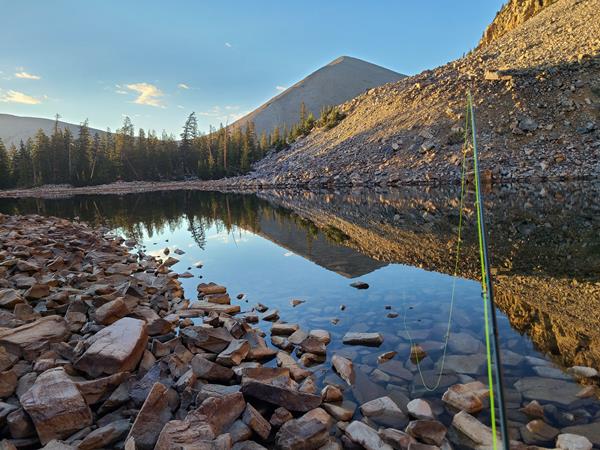 A reflective lake surrounded by rocks, with a green fishing rod emerging from the viewer.
