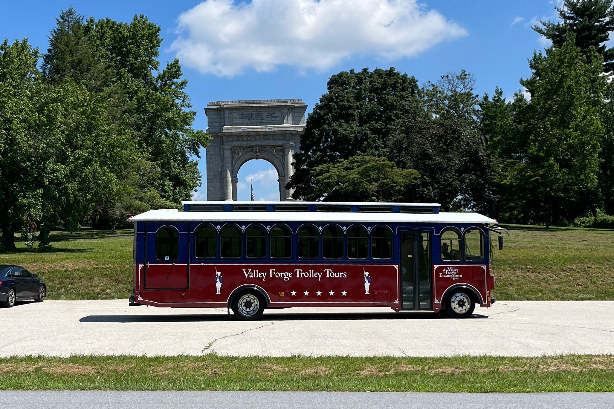 a trolley drives past a large arch monument and trees.