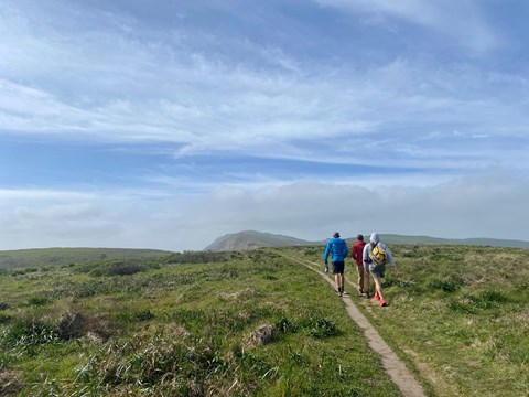 Three hikers on a dirt, single track trail through lush green grass.