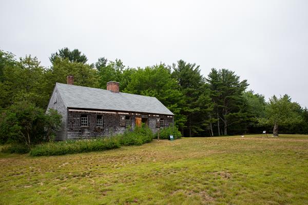 A grey, wood-shingle building with two chimneys sits in a grassy field with trees in the background