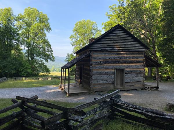 Simple log cabin surrounded by a split-rail fence with a mountain landscape in the background.