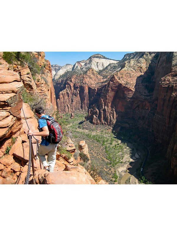 A hiker in a blue shirt holds onto chains as they traverse a sandstone cliff.