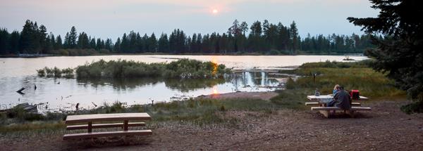 A couple sits at one of two picnic tables on the shore of a lake lined by conifers with the setting