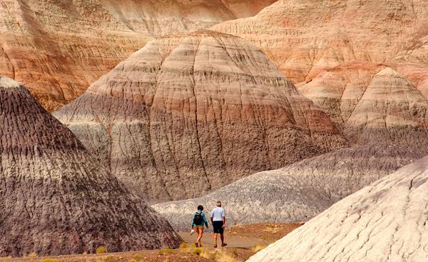 Two hikers walk amidst gray, blue, and purple badlands along a trail