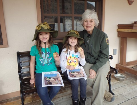 Two kids sit on a bench with a park volunteer. They're holding up badges.