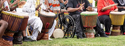 A line of colorfully dressed seated people in colorful clothes beat drums with their hands. .