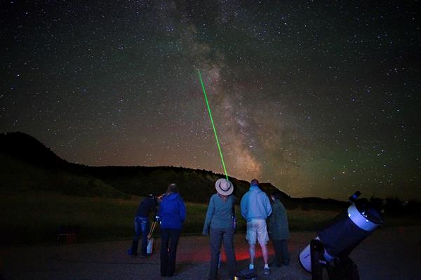 A ranger stands with several people, pointing a green laser at the sky. Stars are visible above them