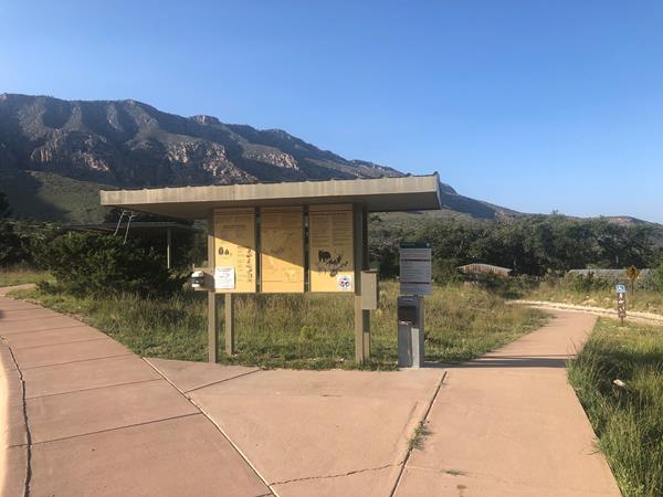 A metal kiosk with trail information stands before a historic ranch compound.
