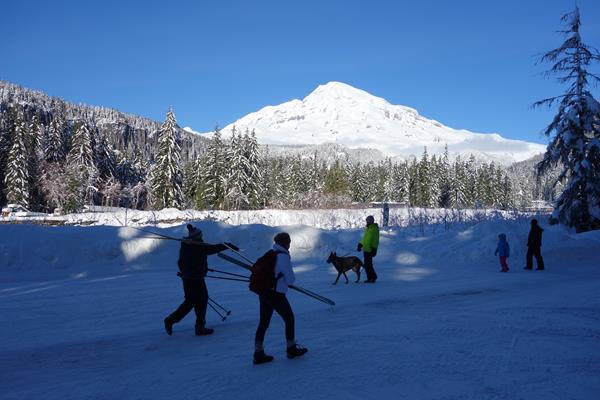 Several people walk along a snowy road in front of a view of a snow-covered mountain and forest.