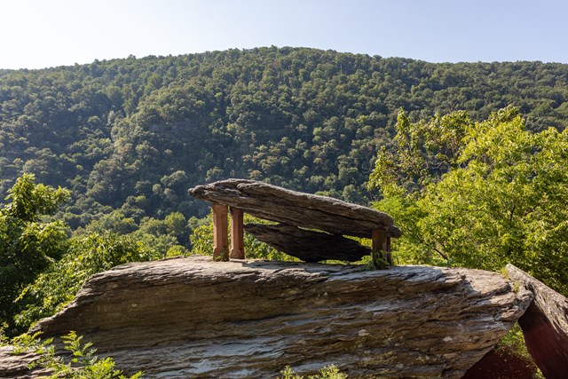 Two flat slabs of slate balanced on red sandstone posts atop a large boulder, with a forested mountain behind
