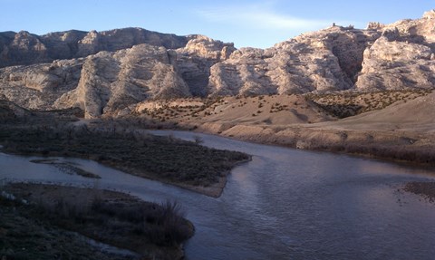 A large river flows in late evening shadow as sun hits a large, rocky mountain in the distance.