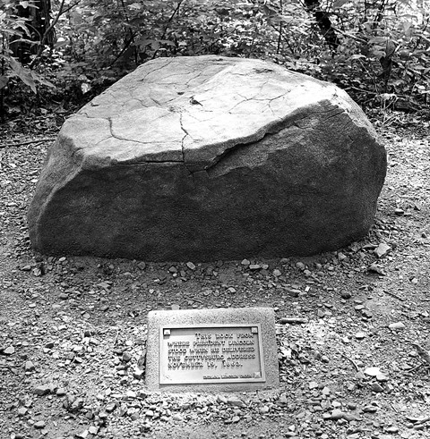 Large stone on side of trail with plaque in front on the ground.