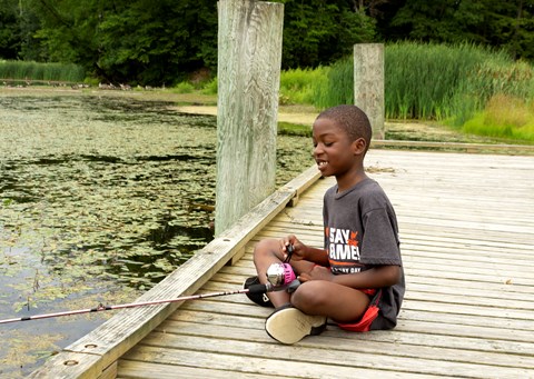A boy sits on the edge of a dock fishing in a pond.