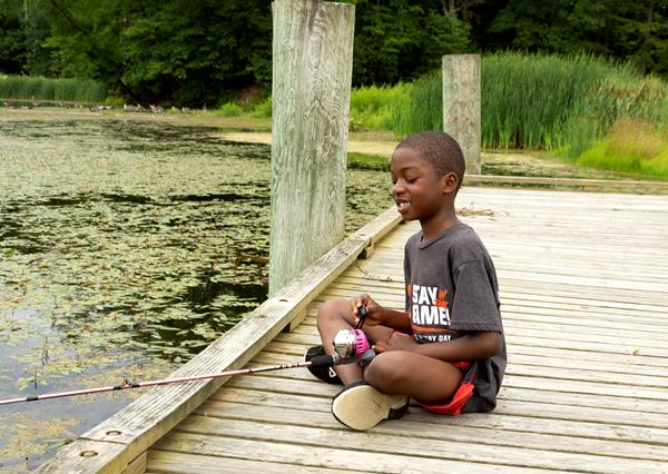 A boy sits on the edge of a dock fishing in a pond.