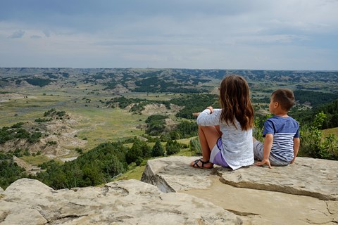 Two children sit on the edge of a rock ledge and gaze out over the badlands, juniper, and trails.