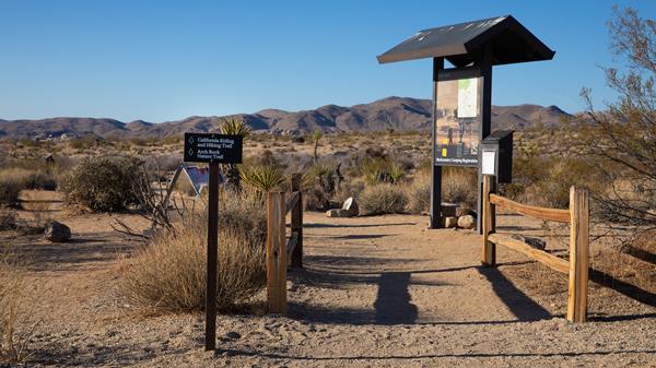 A dirt trail between split rail fences with a backpacking information board surrounded by shrubs.