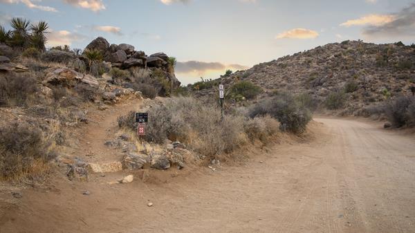 A dirt road going by rocky hills with a dirt trail leaving the road and heading over a rocky hill.