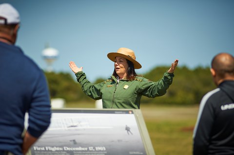 A uniformed park ranger speaks in front of visitors with arms out open.