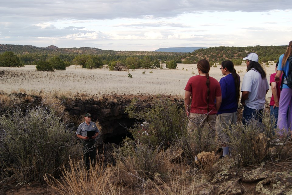 5 people stand watching a park ranger in front of the opening of a cave