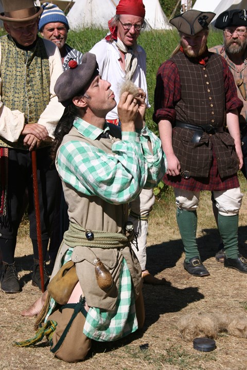 People watching a kneeling man in historic clothing blowing on some fluff.