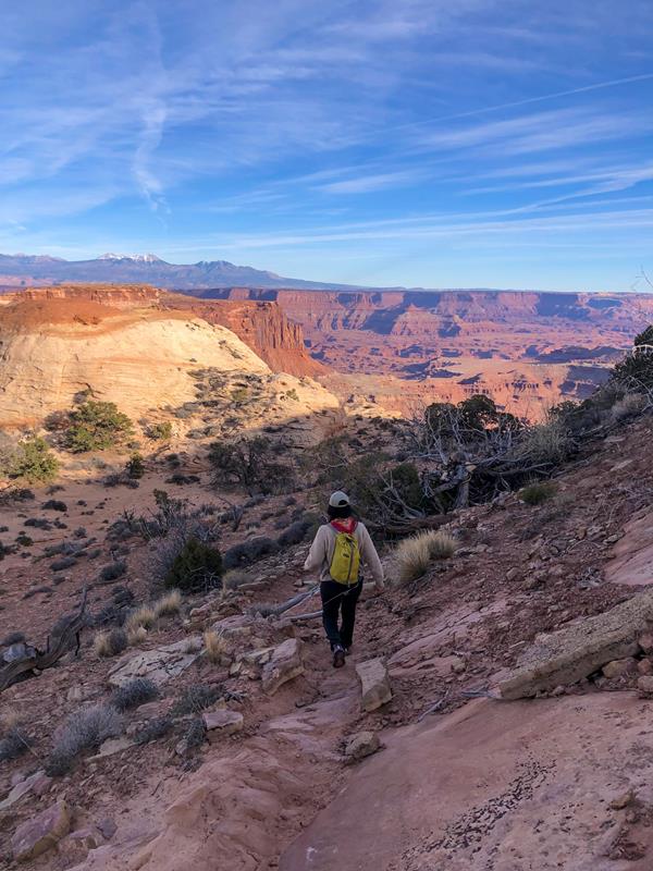 A hiker descends the trail into a canyon, sweeping views are visible in the distance.