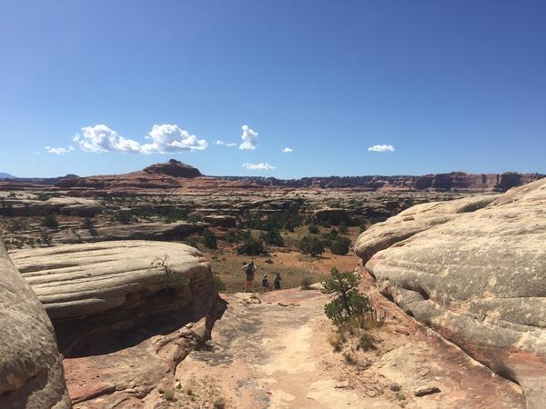 Two hikers are seen in the distance before descending into a canyon on a bright, bluebird day