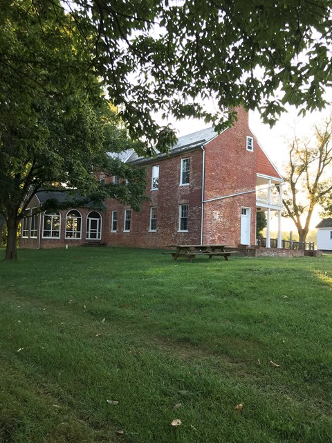 A picnic table sits on a green lawn with a large,P red brick house behind it.