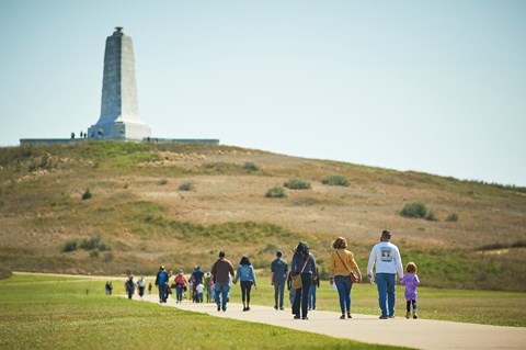 Visitors walk along a sidewalk towards a large green, grass covered hill where the monument sits.