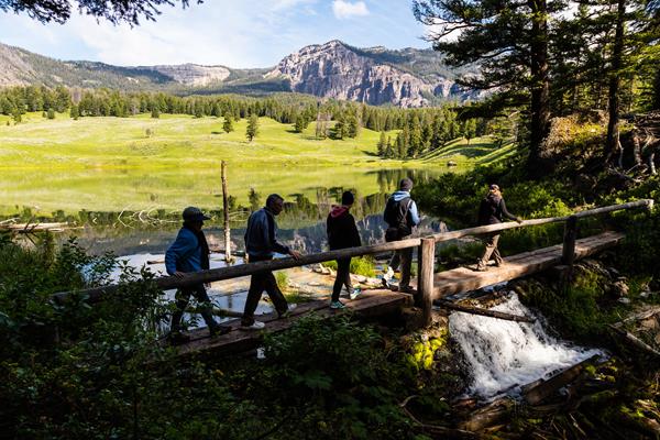 Hikers walk across a bridge at the outlet of a small lake surrounded by rolling hills.
