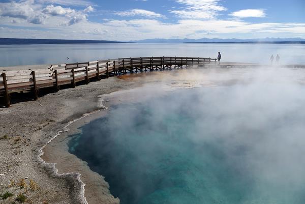 A turquoise hot spring pool surrounded by a boardwalk sits above a large lake.