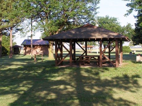 Picnic table under a shelter surrounded by trees with brick restroom in background.