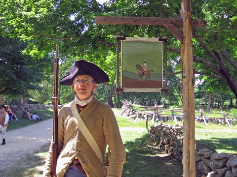 Revolutionary War soldier with a musket standing beneath a painted wooden tavern along a dirt roada