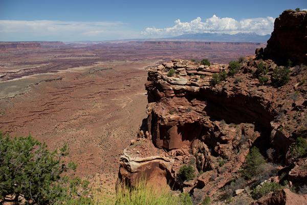 A rocky, steep cliff edge with vegetation in the foreground, far off canyons, mountains in the back.