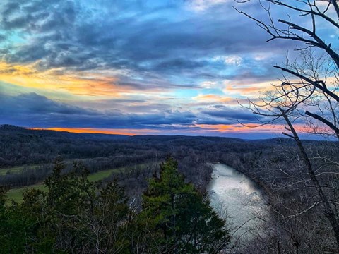 A dusk photo overlooking a river an forest with purple and pink and blue clouds as the sun sets