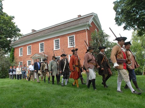 A line of people dressed as 18th C militia and in modern clothes march past a large brick house.