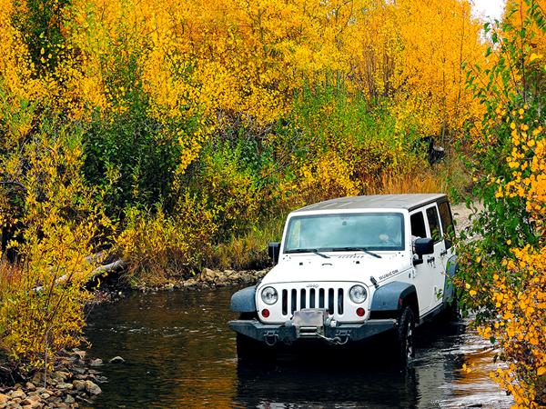 A white Jeep crosses a creek surrounded by gold aspen trees