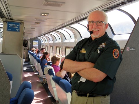 Man in green shirt poses in foreground of an Amtrak sightseer train car.
