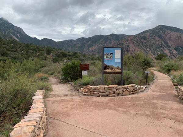 a sidewalk with stonework walls leads toward a metal trailhead sign with mountains in the background