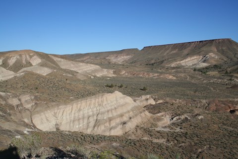 A tan geologic formation sits in a valley with plateaus in the background