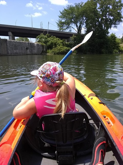 You are seated behind a little girl with a ponytail. You are in a kayak and she is holding a paddle.