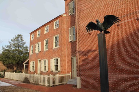 A statue of a raven sitting atop a plinth stands outside a three story brick house.