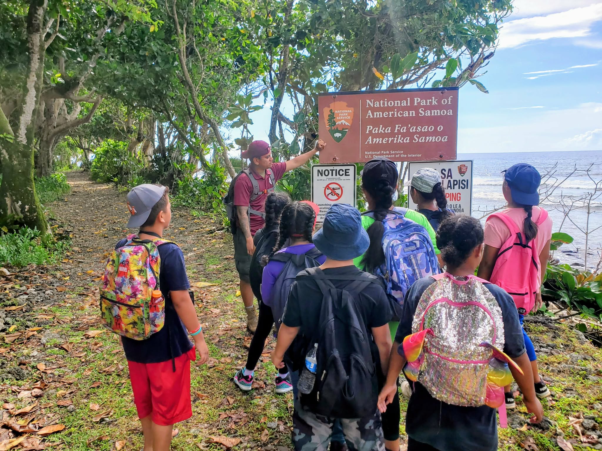 Outdoors; group of young adults facing an adult standing next to sign, trail and forest behind.