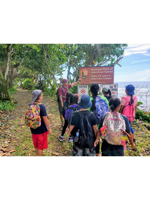 Outdoors; group of young adults facing an adult standing next to sign, trail and forest behind.
