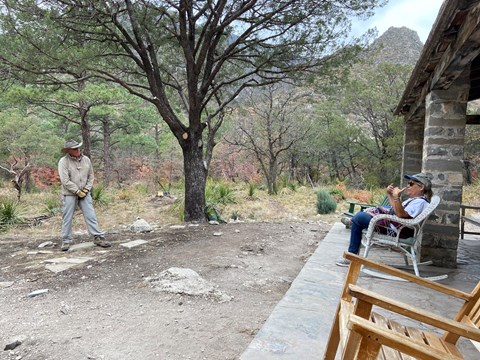 Volunteer speaks to a visitor seated on the porch of a stone cabin