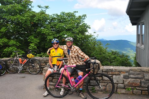 Two bicyclists sit on a stone wall near their bikes. Forest and mountains in background.