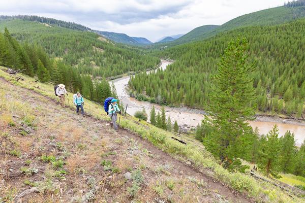 Three backpackers walk along a trail above a forested river valley.