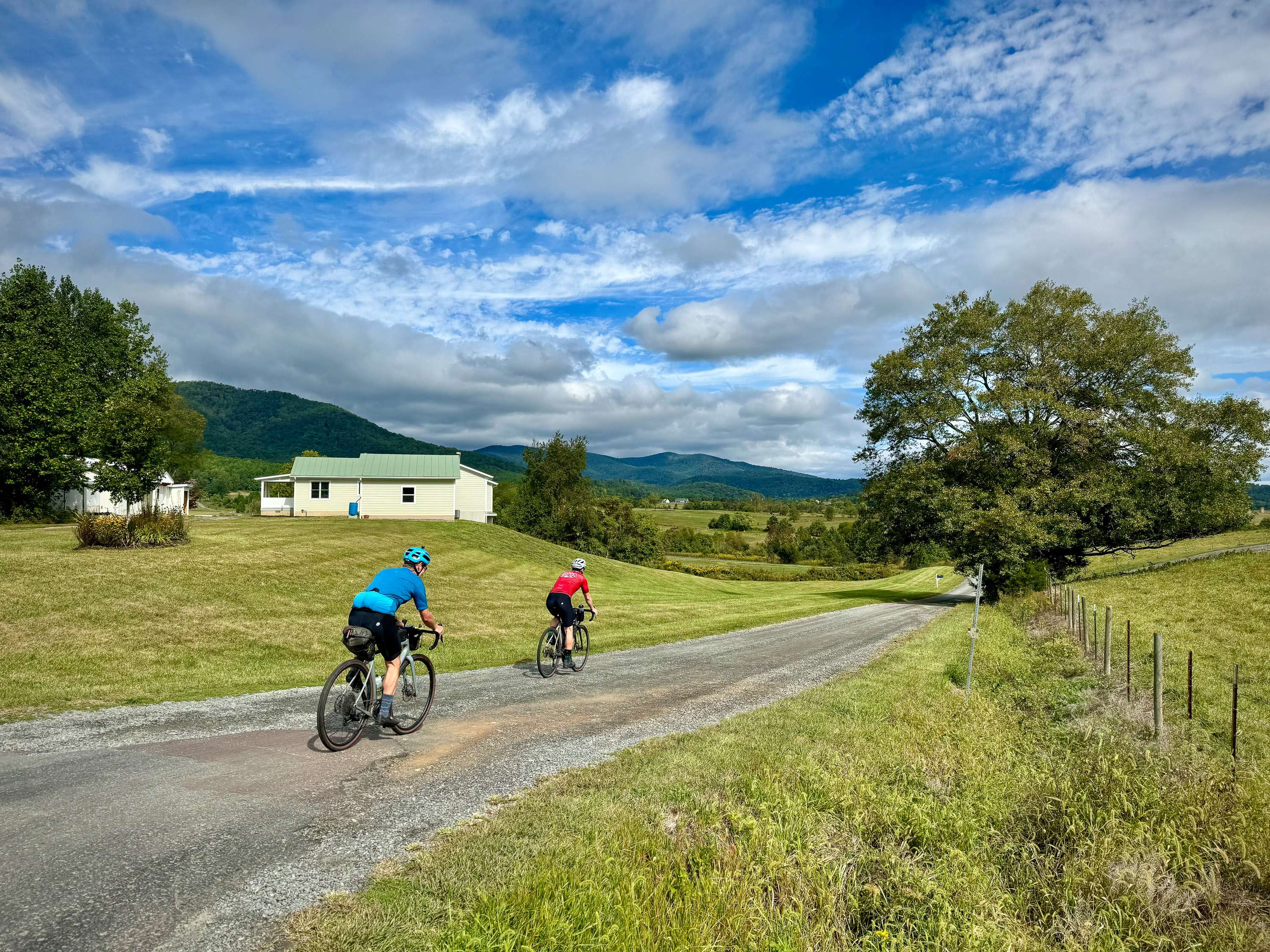 Two cyclists on a gravel road with hills and a house in the background under a cloudy blue sky.