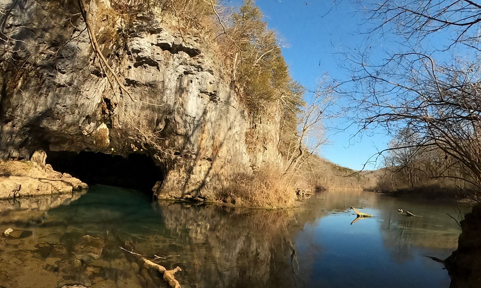 Clear but blueish water with cave opening on left, surrounded by grey rock and a hill