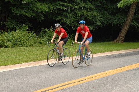 Two bicyclists ride next to each other wearing matching red shirts