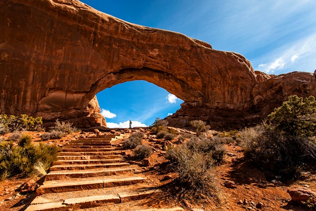 Rock steps lead to an orange stone arch towering over a tiny person standing beneath it